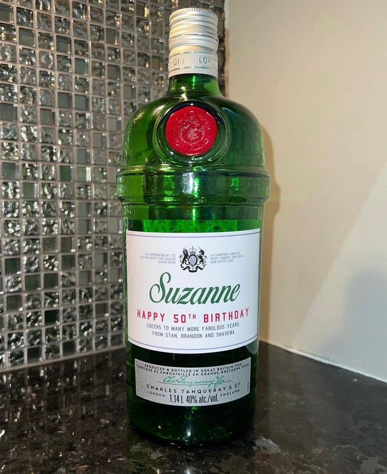May include: A green glass bottle of Tanqueray gin with a personalized label that reads "Suzanne Happy 50th Birthday Cheers to many more fabulous years from Stan, Brandon and Shavena". The label also includes the text "Charles Tanqueray & Co London 1.14L 40% alc./vol. England".