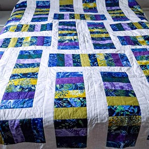 May include: A patchwork quilt with a rectangular block design. The quilt uses blue, purple, and yellow fabrics, separated by white borders. The fabric patterns include floral and abstract designs, creating a vibrant and colourful textile.