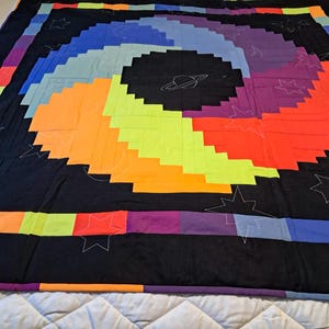 May include: A vibrant quilt with a spiral design, featuring a black background and blocks of neon green, orange, purple, and blue. White embroidered stars, a moon, and a planet decorate the quilt.