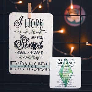 Op de afbeelding: Een witte kaart met zwarte handgeschreven tekst die luidt "Ik werk hard zodat mijn Sims alle uitbreidingspakketten kunnen hebben". Een groen-witte kaart met de tekst "In geval van nood, Ctrl+Shift+C" en een lijst met cheatcodes voor het videospel De Sims.
