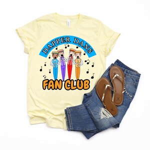 May include: A light yellow t-shirt with the text "Dapper Dans Fan Club" and a graphic of four singing men in colorful outfits. The shirt is paired with blue jeans and brown sandals.