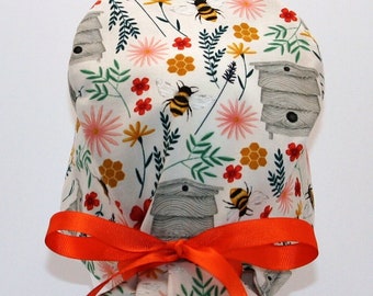 May include: A white scrub hat with a floral print featuring bees, beehives, and honeycomb. The hat is tied with a bright orange ribbon.