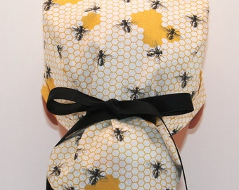 May include: A white and yellow surgical scrub hat with a honeycomb pattern and black bees. The hat has a black tie at the back.