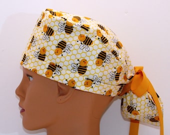 May include: A white scrub hat with a yellow and black bee pattern. The hat has a yellow tie that is tied at the back of the head.