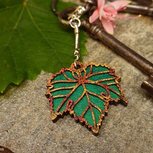 Può includere: Un portachiavi a forma di foglia verde con dettagli dorati e rossi è appeso a una catena d'argento. Il portachiavi è accanto a una foglia verde, chiavi antiche e un fiore rosa. Accessorio decorativo.