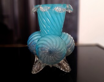 Könnte beinhalten: Eine türkisfarbene Glasvase mit einem Spiralmuster. Die Vase hat eine ausgestellte Oberseite mit einem gerüschten Rand und wird von drei kugelförmigen Basen und klaren Glasfüßen getragen. Das Glas hat eine durchscheinende Qualität.