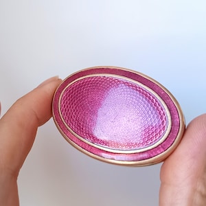 Könnte beinhalten: Eine ovale Brosche mit einem leuchtend magentafarbenen Emaille-Zentrum und einem detaillierten Guilloché-Muster. Das Stück ist von einem glatten, polierten goldfarbenen Metallrand umgeben, der einen Kontrast zwischen der texturierten Oberfläche bildet.
