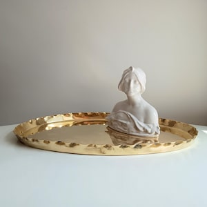 Puede incluir: Una bandeja ovalada dorada con borde festoneado. Un busto de cerámica blanca de una mujer descansa sobre la bandeja. La bandeja está sobre una superficie blanca, con un fondo neutro.