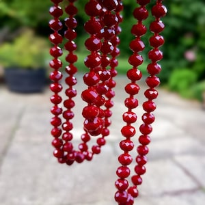May include: Close-up of multiple strands of faceted, deep red beads. The beads are strung together, creating a necklace-like appearance. The beads have a glossy finish and reflect light. The background is blurred green.