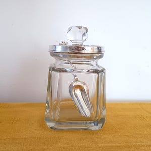 Puede incluir: Un tarro de cristal transparente con borde plateado y tapón de cristal facetado. Una pequeña cuchara plateada descansa dentro del tarro. El tarro es cuadrado con bordes biselados. El tarro está sobre una superficie amarilla.