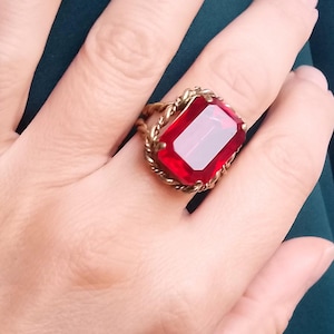 Pode incluir: Um anel de ouro com uma grande gema retangular vermelha engastada em uma banda torcida.