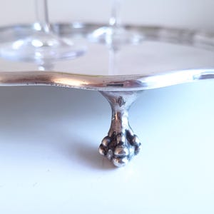 Op de afbeelding: Een zilverkleurig metalen serveerblad met een glad, reflecterend oppervlak en een geschulpte rand. Het blad wordt ondersteund door drie opvallende pootjes in klauw-en-balstijl, met een klein keurmerk zichtbaar op de poot.