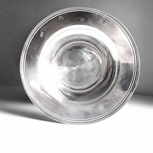 May include: A circular silver bowl with a polished, reflective surface and a wide, flat rim. The rim features four distinct stamped hallmarks evenly spaced along the top edge, indicating metal purity and origin. The bowl has a deep, concave centre.