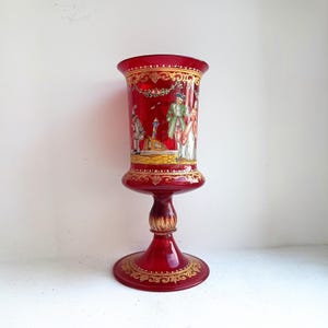 May include: A tall, ruby-red glass goblet with a wide rim and a decorative base. The goblet features a painted scene of figures in period clothing, accented with gold filigree. The stem is a dark, ornate gold colour.
