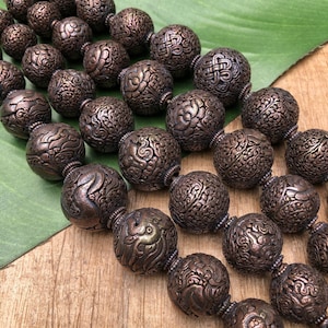 Peut inclure: Un collier de perles rondes brunes avec des motifs gravés complexes. Les perles sont en métal et ont une finition légèrement brillante.