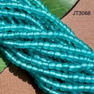 May include: A strand of turquoise blue glass beads. The beads are small and round, with a frosted finish. The beads are arranged in a loose pile, and the image is taken from a close-up perspective. JT3068