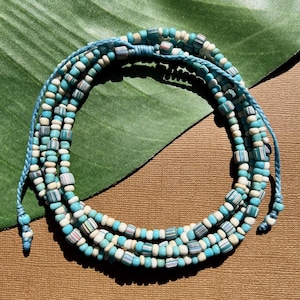 May include: A multi-strand bracelet featuring turquoise, white, and striped beads. The bracelet is strung on a light blue cord, with adjustable ties at the ends. The beads are a mix of round and rectangular shapes, creating a textured look.