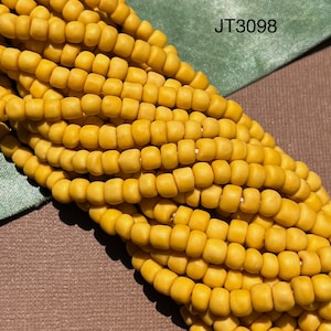Pode incluir: Um close-up de um fio de contas de sementes amarelas. As contas são pequenas e redondas, e estão dispostas em um padrão apertado e uniforme. As contas são enfiadas em um fio fino e branco. A imagem é rotulada como "JT3098".