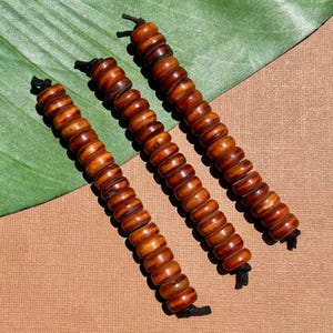 May include: Three strands of brown wooden beads, each strung on black cord. The beads are cylindrical with rounded edges, displaying variations in color from light to dark brown. The beads are arranged diagonally against a green leaf and a brown surface.