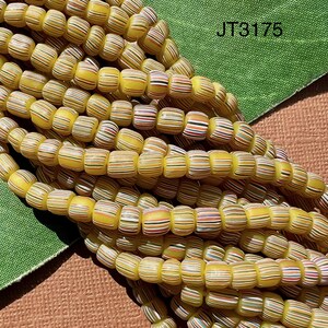 May include: A close-up of a strand of yellow and white striped glass beads. The beads are arranged in a random pattern and are all the same size. The beads are on a green and brown surface. JT3175