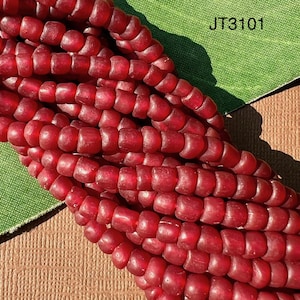 Pode incluir: Close-up de um fio de contas de vidro vermelhas. As contas são pequenas, cilíndricas e têm uma aparência ligeiramente translúcida. As contas são enfiadas, formando uma superfície densa e texturizada. O texto "JT3101" é visível no canto superior direito.