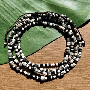 May include: A multi-strand beaded necklace with black, white, and striped beads. The beads are strung on a brown cord, creating a layered effect. The necklace is displayed on a green leaf and a brown surface.