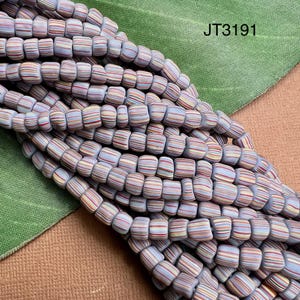 Może przedstawiać: Zbliżenie sznura kolorowych, pasiastych koralików. Koraliki są cylindryczne, z jasnoniebieską podstawą i poziomymi paskami w różnych kolorach, w tym czerwonym, żółtym i brązowym. Widoczny jest tekst "JT3191".