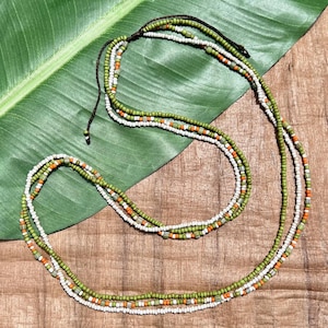 May include: A layered beaded necklace with green, white, and orange beads. The necklace is displayed on a green leaf and a wooden surface. The necklace has a dark brown cord for adjusting the length.