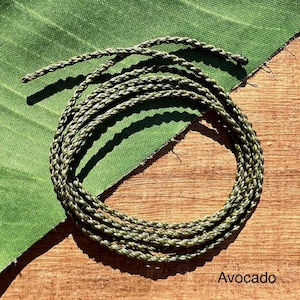 May include: Coiled green and tan rope, labeled "Avocado", rests on a wooden surface next to a green fabric. The rope is tightly braided, showcasing a mix of green and tan hues. The background features a textured green fabric.