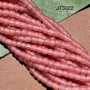 Pode incluir: Um fio de contas de vidro rosa, com acabamento fosco. As contas são pequenas e redondas, e são enfiadas em um fio transparente. As contas são dispostas em um padrão solto e fluido. A imagem inclui o texto "JT3022".