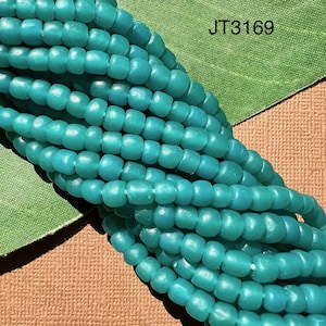 Può includere: Un filo di perline di vetro blu turchese. Le perline sono piccole e rotonde, con una finitura leggermente opaca. Le perline sono infilate su un filo trasparente. L'immagine include il testo "JT3169".