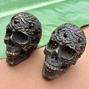 May include: Two antique brass skull figurines with intricate floral patterns. The skulls have open eye sockets, nasal cavities, and mouths. The skulls are displayed on a brown surface with a green leaf in the background.