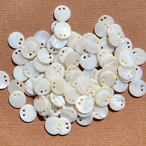 May include: A collection of numerous small, round, white mother-of-pearl buttons. Each button features a smooth, iridescent surface with two small holes positioned near the top edge for sewing. The buttons are scattered in a dense, overlapping pile.