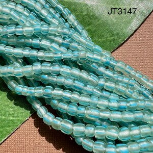 May include: A strand of light blue glass beads with white stripes. The beads are opaque and have a slightly frosted finish. The beads are arranged in a loose pile, and the image is taken from a close-up perspective. JT3147