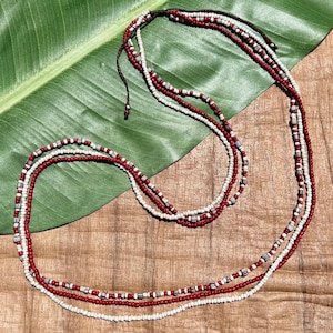 以下が含まれることがあります： 赤、白、グレーのビーズを使った多層ネックレス。 ネックレスは、小さくて丸いビーズを複数連ねて作られており、質感とディテールが際立っています。 ビーズは濃い茶色のコードに通されており、木製の表面に展示されています。