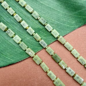 May include: Close-up of three strands of light green beads. Each rectangular bead has a carved design with small black circles. The beads are strung together with smaller, round beads. The background is a green fabric.