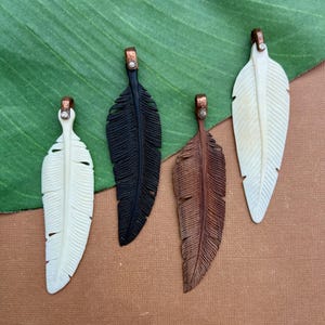 Puede incluir: Cuatro colgantes de plumas en tonos marfil, negro y marrón. Cada colgante presenta un diseño de pluma tallada con una argolla de color cobre. Los colgantes se exhiben sobre una hoja verde y una superficie marrón.