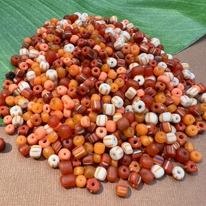 May include: A large pile of small, colorful glass beads in shades of orange, red, and white. The beads are mostly round and have a variety of patterns, including stripes and dots.