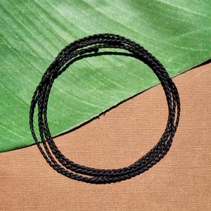 May include: A coil of black cord lies on a green leaf and a brown surface. The cord is tightly woven, and the circular shape is centered in the frame. The background is a mix of green and brown.