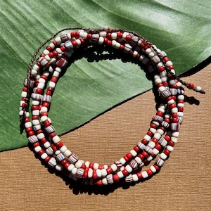 Puede incluir: Un collar de cuentas de varias hebras con cuentas rojas, blancas y rayadas. El collar está ensartado en un cordón marrón y se exhibe sobre una superficie marrón con una hoja verde en el fondo. Las cuentas son pequeñas y cilíndricas.