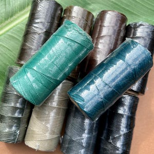 May include: A collection of spools of colorful waxed cord in shades of green, brown, and blue. The spools are wrapped in clear plastic, and the cord appears to be tightly wound. The cords are likely used for jewelry making or other crafts.