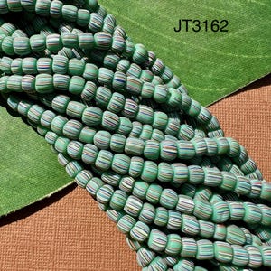May include: A strand of green, white, and red striped glass beads. The beads are small and round, and they are strung together on a clear thread. The beads are arranged in a pile, and they are lit from above. The image is labeled "JT3162".