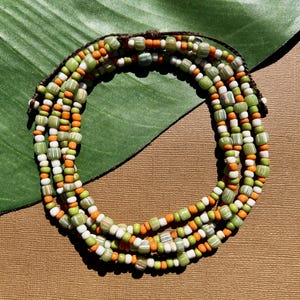 May include: A multi-strand beaded necklace with green, orange, and white beads. The beads are strung on a brown cord, creating a layered effect. The necklace is displayed on a brown surface with a green leaf in the background.
