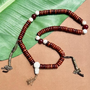 Puede incluir: Un rosario de cuentas de madera marrón oscuro, intercaladas con cuentas blancas y granates. Las cuentas están ensartadas en un cordón marrón y tienen detalles metálicos y borlas.