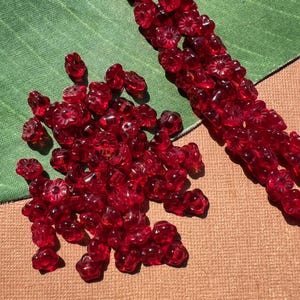 Könnte beinhalten: Eine Sammlung von durchscheinenden roten Glasperlen in Blumenform. Einige Perlen sind verstreut, andere sind in einer Reihe aufgereiht. Die Perlen liegen auf einer grünen und beigen Oberfläche.