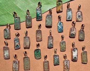 Puede incluir: Una colección de 20 colgantes de vidrio antiguo en varios tonos de verde, azul y marrón. Cada colgante es rectangular y tiene una anilla de metal dorado. Los colgantes están dispuestos sobre una superficie marrón.