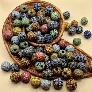 Può includere: Una collezione di perline di vetro colorate e decorate in un cestino di vimini. Le perline presentano vari disegni, tra cui motivi floreali, geometrici e astratti in rosso, blu, giallo e verde. Le perline sono rotonde e hanno un foro al centro.
