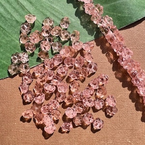 Può includere: Perline rosa a forma di fiore sparse su una superficie marrone, con un filo di perline visibile. Le perline traslucide hanno un delicato design floreale, perfette per la creazione di gioielli o progetti artigianali.