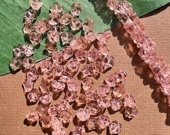 May include: Pink flower-shaped beads scattered on a brown surface, with a strand of beads visible. The beads are translucent and have a delicate, floral design, perfect for jewelry making or crafting projects.