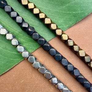 Puede incluir: Primer plano de tres hilos de cuentas metálicas. Las cuentas tienen forma de cubo con bordes redondeados. Un hilo es plateado, otro gris oscuro y el tercero dorado. Las cuentas están dispuestas en diagonal sobre un fondo verde y marrón.